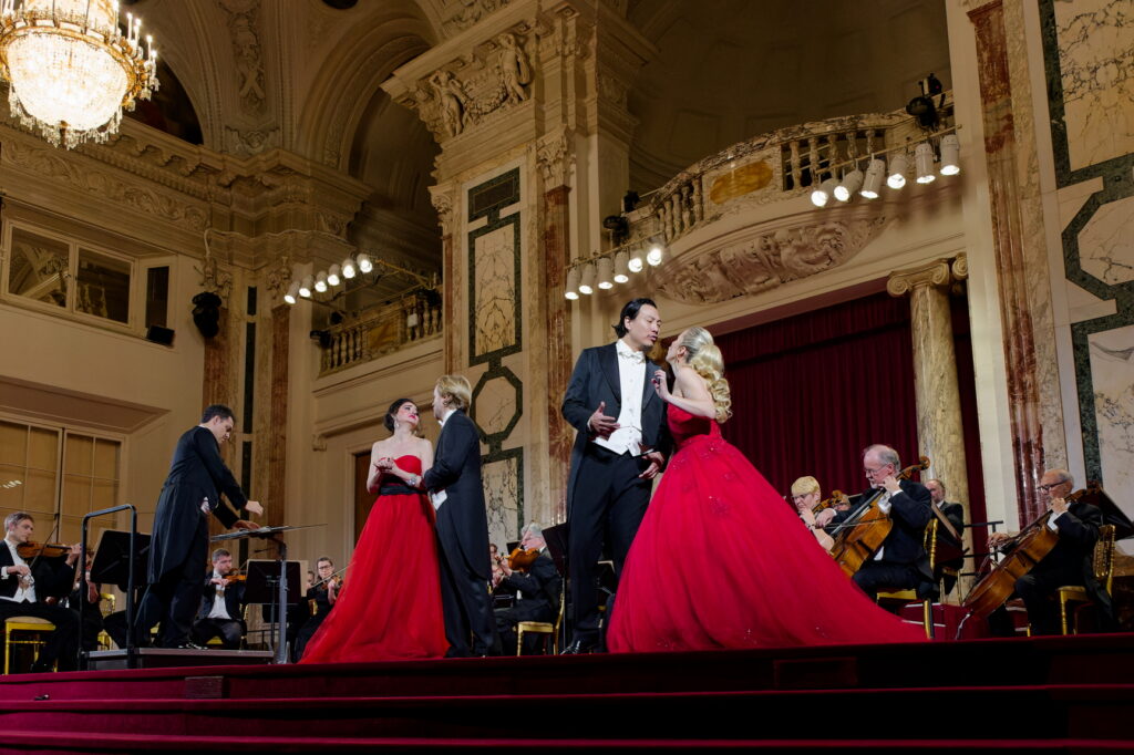 Opernsänger in festlicher Kleidung bei einer Aufführung, begleitet von einem Orchester in einem prunkvollen Saal