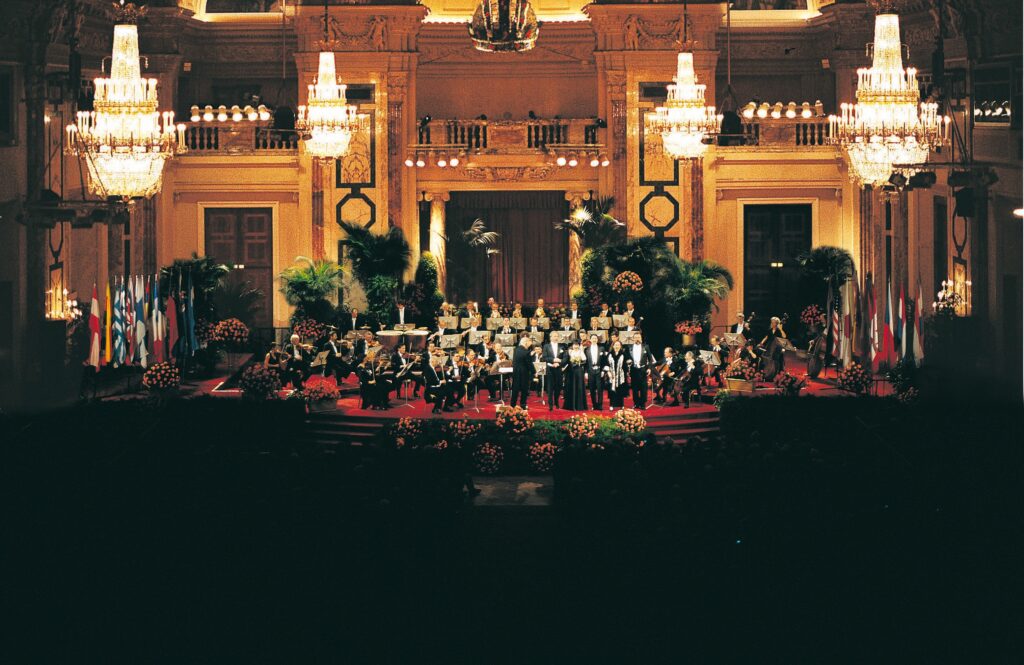 Prunkvoller Festsaal mit Kronleuchtern, Orchester auf der Bühne, dekoriert mit Fahnen und Pflanzen